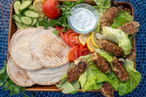 Dinner spread with pita bread, lettuce, tomato, cucumber, yogurt sauce and chicken kofta. On a blue table.