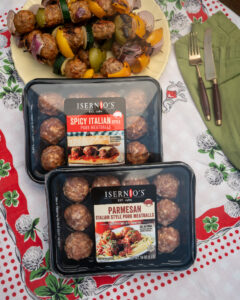 Packaged Isernio's meatballs on a table. A tray of meatball and vegetable kebabs behind the tray.