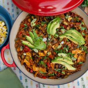 Red skillet with ground chicken enchilada recipe topped with melted cheese and sliced avocado. A bowl of corn is served on the side.