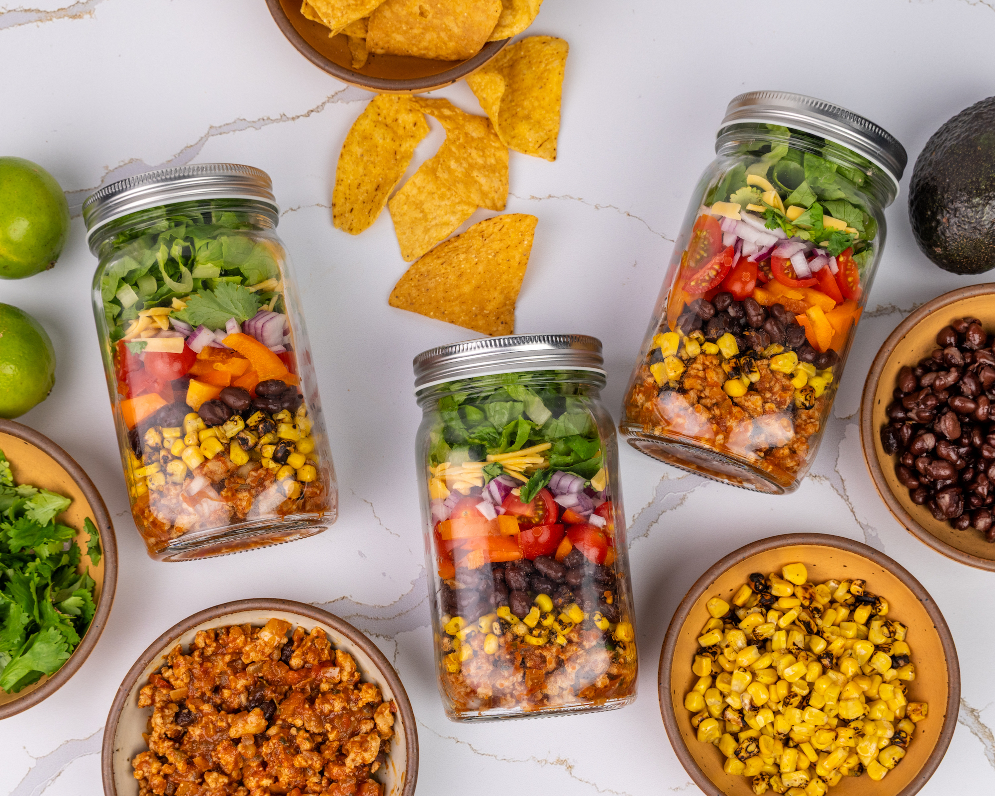 A meal prep photo of three large mason jars filled with layers of seasoned ground chicken, beans, corn, lettuce, cheese and tomatoes.