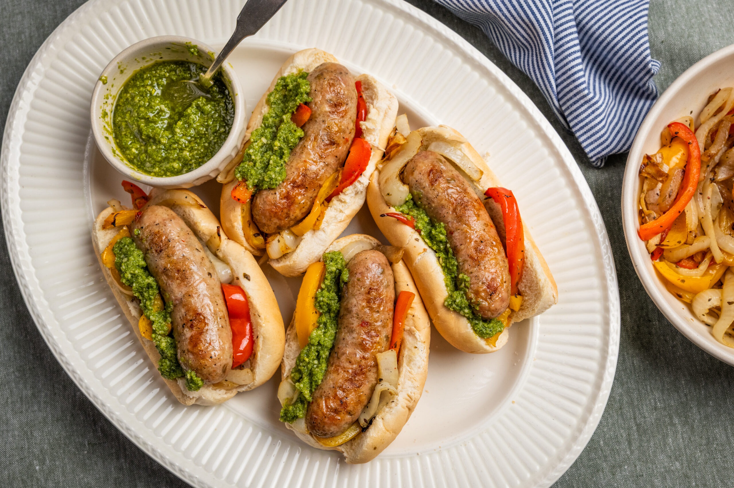 Grilled Isernio’s Mild Italian Chicken Sausage sandwiches topped with fresh basil pesto and sautéed bell peppers and onions, served on soft sandwich rolls on a white platter. A bowl of homemade pesto and extra grilled peppers and onions accompany this easy summer grilling recipe, perfect for backyard cookouts, Father’s Day celebrations, and family dinners.