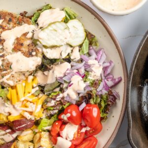 Overhead view of a Chicken Burger Bowl made with Isernio's Ground Chicken. A seasoned chicken burger patty is served over fresh mixed greens with roasted sweet potatoes, sliced avocado, shredded cheddar cheese, cherry tomatoes, diced red onion, and pickle slices, all drizzled with a creamy burger sauce. Additional chicken burger patties cook in a cast-iron skillet alongside the finished bowl.